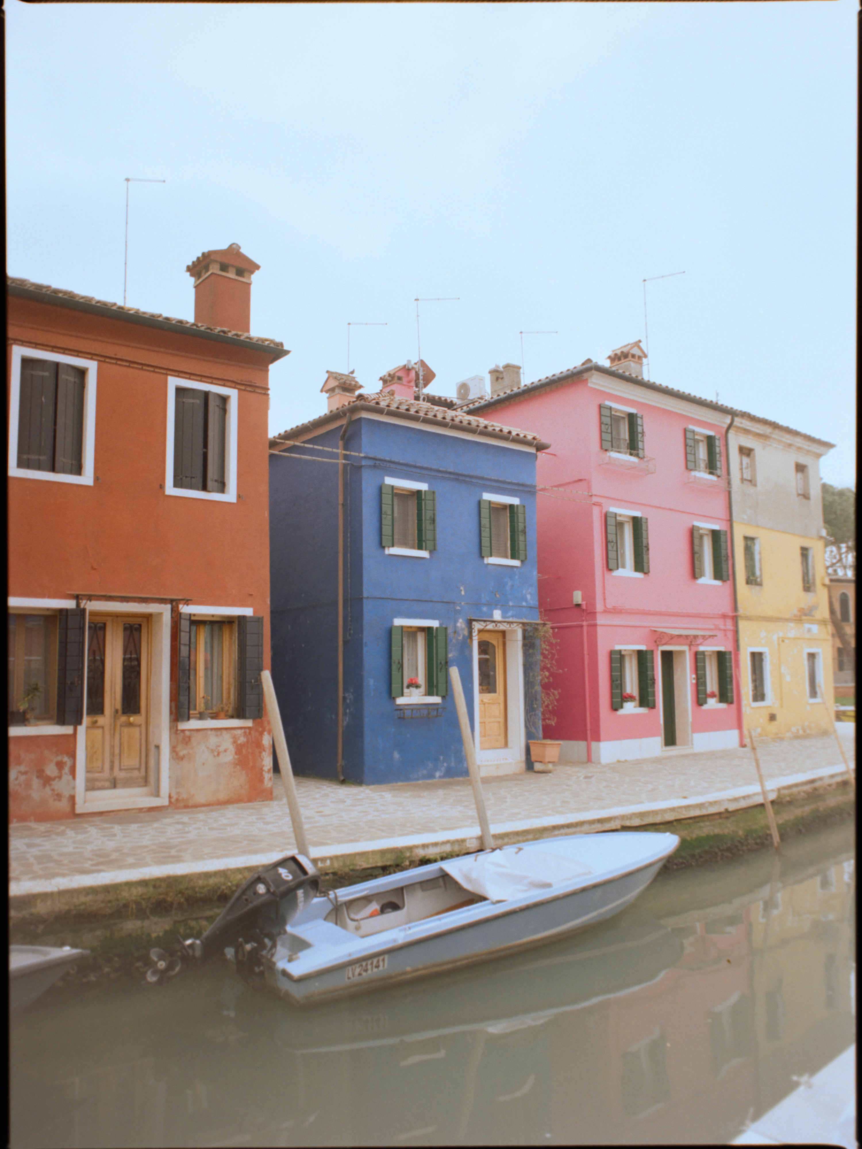 Four canalside houses in orange, navy, pink and yellow with a white boat — Burano colour blocking on film